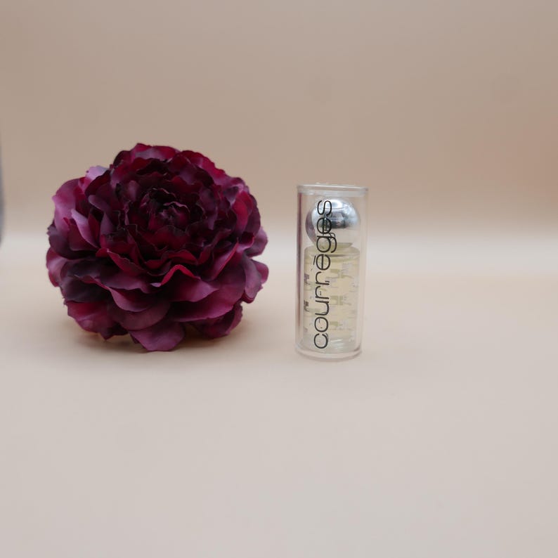 May include: A clear perfume bottle with a silver cap and the word "courr&egrave;ges" printed vertically. A large, dark burgundy flower is to the left of the bottle. The background is a solid, light beige color.