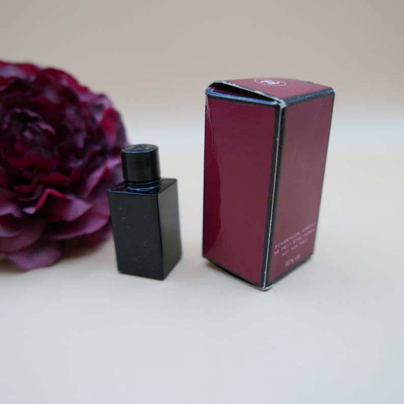 May include: A small, black perfume bottle with a matching cap is next to a burgundy box. A dark red flower is in the background. The bottle has a square shape and a simple design.