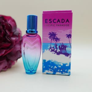 May include: A glass perfume bottle with a blue cap and a pink-to-blue gradient body next to its rectangular box. The box features the text ESCADA PACIFIC PARADISE, a beach scene with palm trees and people, and the volume 0.14 FL OZ 4 ml.