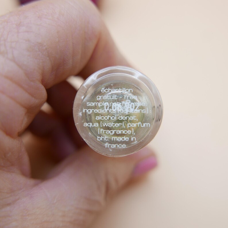 May include: A clear glass bottle with a silver cap, held in a hand. The bottle contains a liquid and has text that reads "&eacute;chantillon gratuit - free sample". The ingredients include alcohol denat, aqua, parfum, and bht. Made in France.