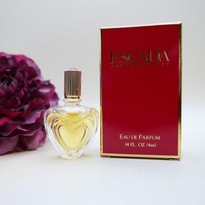 May include: A heart-shaped glass perfume bottle with a gold cap containing yellow liquid, positioned next to a rectangular red box with gold trim. The box features the text ESCADA MARGARETHA LEY EAU DE PARFUM .14 FL. OZ./4ml in gold lettering.