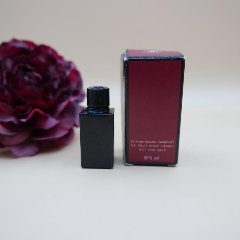 May include: A small, black perfume bottle with a matching cap is next to a burgundy box. The box has text that reads "ECHANTILLON GRATUIT NE PEUT ETRE VENDU NOT FOR SALE 80% vol". A dark red flower is in the background.