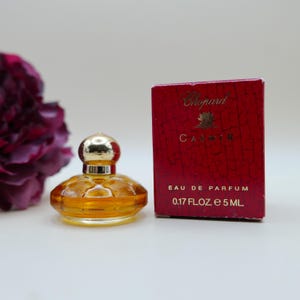 May include: A miniature Chopard Casmir eau de parfum bottle with a gold-toned cap and amber liquid, positioned next to its matching red box featuring a cracked texture, gold lettering, and the printed volume of 0.17 fl. oz. and 5 ml.