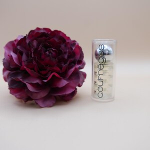 May include: A deep burgundy artificial flower next to a clear Courr&egrave;ges perfume bottle. The bottle has a silver cap and the Courr&egrave;ges logo in white. The flower has multiple layers of petals.