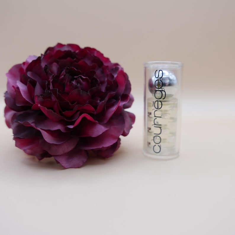 May include: A deep burgundy artificial flower next to a clear Courr&egrave;ges perfume bottle. The bottle has a silver cap and the Courr&egrave;ges logo in white. The flower has multiple layers of petals.