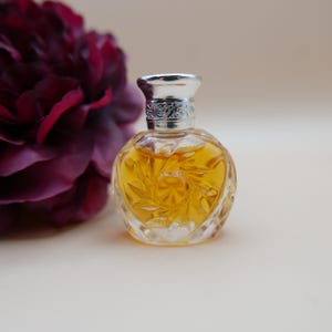 May include: A small, round, clear glass perfume bottle with a faceted, starburst-like pattern on the body. The bottle is filled with an amber-coloured liquid and features a silver-toned metal cap with an engraved, decorative band around the neck.