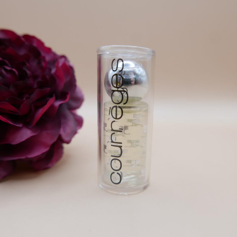 May include: A clear Courr&egrave;ges perfume bottle with a silver cap is encased in a transparent tube. The bottle is filled with a light-colored liquid. The Courr&egrave;ges logo is printed vertically on the bottle. A dark red flower is in the background.