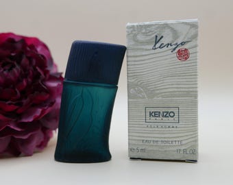 Può includere: Un flacone in miniatura di Eau de Toilette Kenzo Pour Homme da 5 ml con contenitore in vetro blu scuro dalla texture a bambù e tappo abbinato, accanto alla scatola rettangolare bianca con motivi a venature del legno e logo Kenzo.