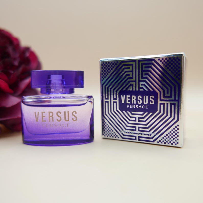 May include: A purple glass perfume bottle with the text VERSUS VERSACE printed on the front, accompanied by a matching square cardboard box featuring a geometric silver and blue pattern and the text VERSUS VERSACE and Eau de Toilette.