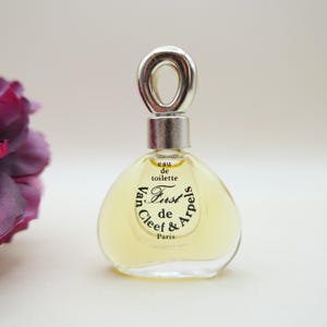 May include: A clear glass perfume bottle containing a pale yellow liquid. The bottle features a silver-toned cap with a circular loop top. Black text on the front reads: eau de toilette, First, Van Cleef & Arpels, Paris.