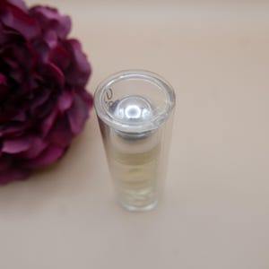 May include: A clear glass perfume bottle with a silver cap, containing a light yellow liquid. The bottle is cylindrical and has a rounded top. A dark purple flower is in the background.