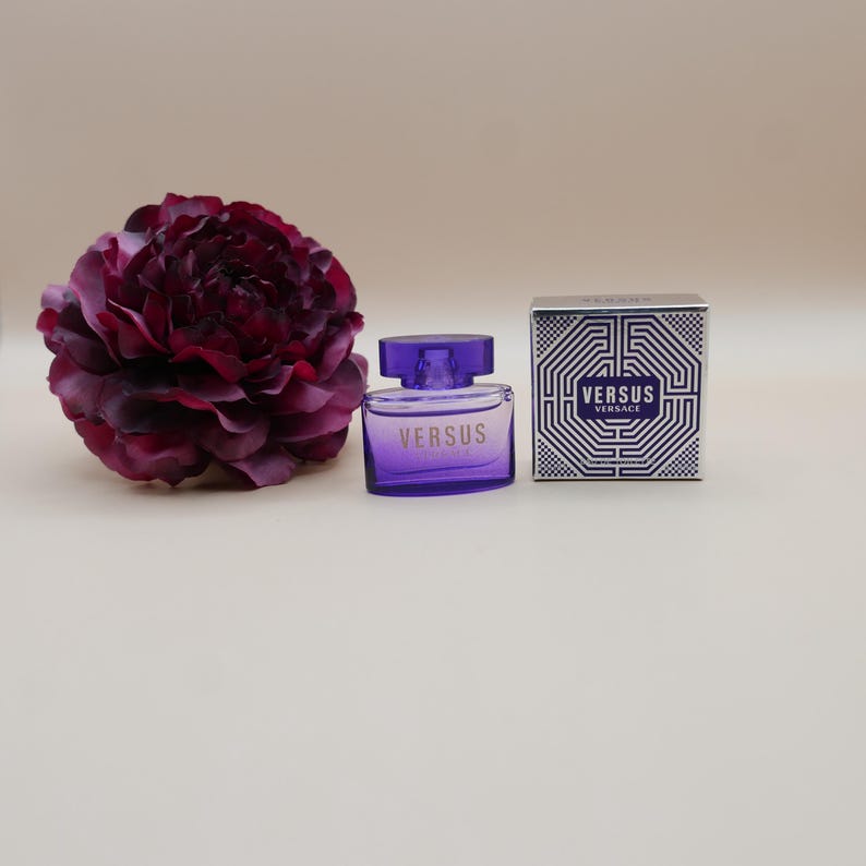 May include: A purple glass perfume bottle with the word VERSUS printed on the front, accompanied by a square box featuring a white and blue geometric pattern and the text VERSUS VERSACE, positioned next to a large, dark purple flower.