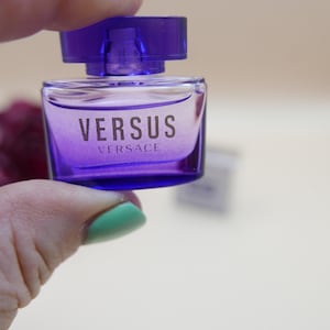 May include: A small, rectangular purple glass perfume bottle with a matching purple cap. The front of the bottle features the text VERSUS VERSACE printed in a dark font. The bottle has a clear, transparent design and a wide, flat shape.
