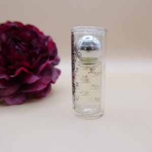 May include: A clear perfume bottle with a silver cap inside a clear plastic tube. The bottle has a silver cap and a clear liquid inside. A dark red flower is in the background.