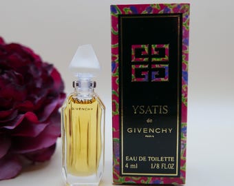 Puede incluir: Un frasco de vidrio en miniatura de Ysatis de Givenchy Eau de Toilette con tapón blanco y líquido dorado. A su lado, una caja rectangular con panel central negro, texto dorado y un borde floral vibrante en tonos rosa, morado y verde.
