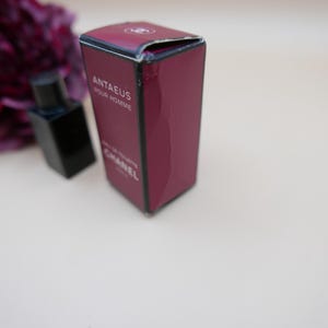 May include: A burgundy box of Chanel Antaeus Pour Homme Eau de Toilette. The box has white text and a black perfume bottle is next to it. The background is a light beige.