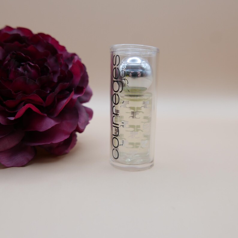 May include: A clear Courreges perfume bottle with a silver cap inside a clear tube. The bottle is next to a dark burgundy flower. The Courreges logo is printed vertically on the tube.