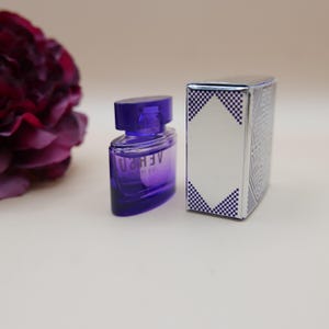 May include: A small purple glass perfume bottle with a matching cap and the text VERSUS printed on the front, positioned next to a rectangular silver box featuring a white diamond-shaped center and blue and white checkered corner accents.