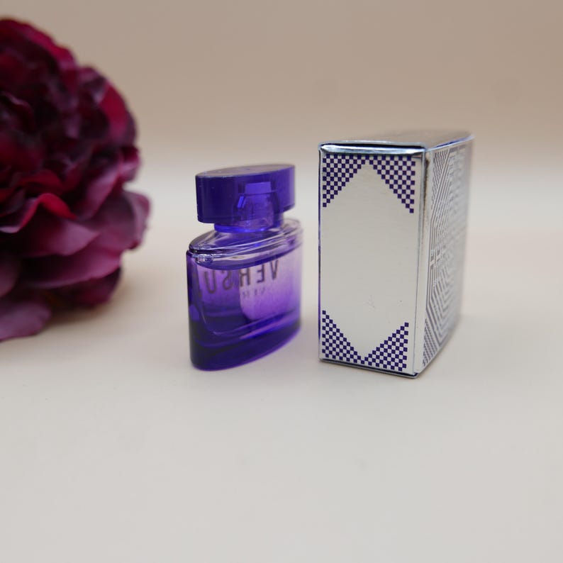 May include: A small purple glass perfume bottle with a matching cap and the text VERSUS printed on the front, positioned next to a rectangular silver box featuring a white diamond-shaped center and blue and white checkered corner accents.
