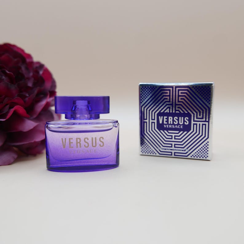 May include: A purple glass perfume bottle with the text VERSUS VERSACE printed on the front, accompanied by a matching square cardboard box featuring a blue and silver geometric maze pattern and the text VERSUS VERSACE in a central rectangle.
