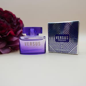 May include: A purple glass perfume bottle with a matching cap and the text VERSUS VERSACE printed on the front, positioned next to a square silver and blue box featuring the text VERSUS VERSACE and a geometric pattern on the front surface.