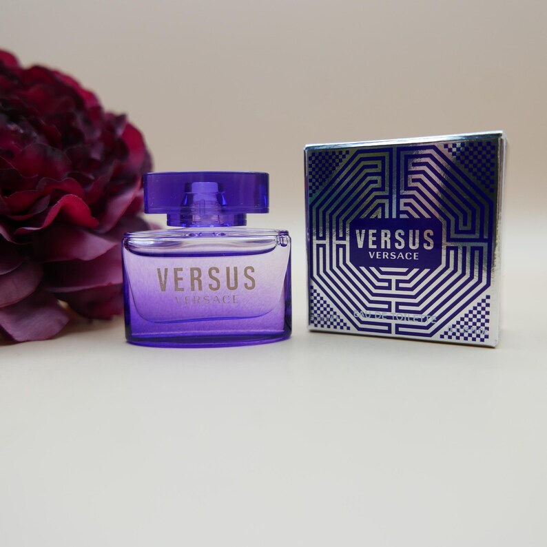 May include: A purple glass perfume bottle with a matching cap and the text VERSUS VERSACE printed on the front, positioned next to a square silver and blue box featuring the text VERSUS VERSACE and a geometric pattern on the front surface.