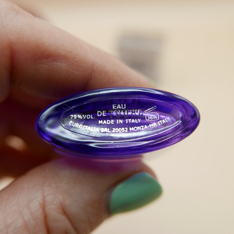 May include: The oval base of a purple glass perfume bottle features white printed text reading EAU DE TOILETTE, 79% VOL., MADE IN ITALY, and EUROITALIA SRL 20052 MONZA-MB ITALY, along with a small icon of an open jar with the text 36M inside.
