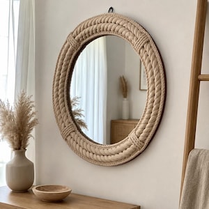 May include: A round wall mirror featuring a thick, beige rope frame wrapped in a braided pattern. The mirror is suspended by a small black hook at the top. The rope texture is prominent, creating a circular border around the reflective glass surface.
