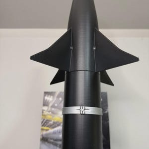 May include: A black and silver model rocket with triangular fins and a conical nose. The rocket has two silver bands around the cylindrical body. The top of the rocket is white.