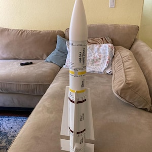 May include: A model missile with a white conical nose and a cylindrical body. The body is primarily white with black, yellow, and brown details, and the text "AIM-54A SERNO 20553" is printed on the side. The model has white fins at the base.