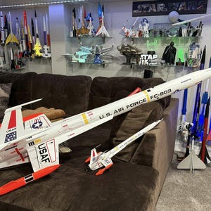 May include: A collection of model rockets and aircraft are displayed on shelves and a brown sofa. The main focus is on a large white model aircraft with red and black accents, and the text "U.S. AIR FORCE FC-803". A smaller model aircraft is also present.