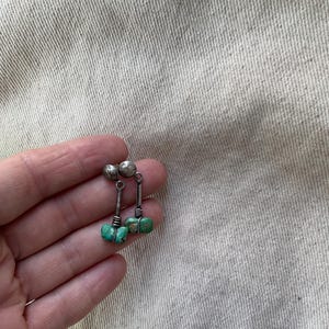 Puede incluir: Pendientes colgantes plateados con detalles de piedras turquesas. Cada pendiente presenta una pequeña bola plateada en la parte superior, un fino poste plateado y un grupo de piedras turquesas en la parte inferior. Fondo de tela neutra.