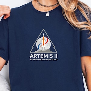 May include: Navy blue t-shirt featuring a triangular graphic with a rocket, planet, and the text "ARTEMIS II TO THE MOON AND BEYOND." The design includes shades of orange, white, and blue. The shirt is a crew neck style.