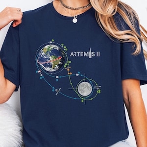 May include: Navy blue t-shirt featuring a graphic of Earth, the Moon, and a spacecraft trajectory, with the text "ARTEMIS II" in white. The design includes colorful dots and lines representing the mission's path. The shirt is a short-sleeved crew neck.