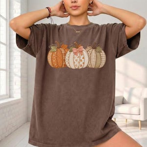 May include: A brown short-sleeve t-shirt featuring a graphic print of three pumpkins arranged in a row. The pumpkins display various patterns including plaid, floral, and solid textures, with a small pink bow attached to the center pumpkin.