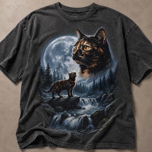 May include: A charcoal gray short-sleeve t-shirt featuring a graphic print of a tortoiseshell cat standing on a rock near a waterfall. Above the cat, a large, detailed moon shines over a dark forest landscape, with a second, larger cat face visible.