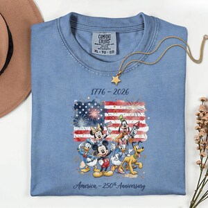 May include: A light blue Comfort Colors t-shirt featuring a graphic of the American flag with fireworks and Disney characters. The text reads "1776-2026" and "America - 250th Anniversary". A gold star pendant necklace rests on the shirt.