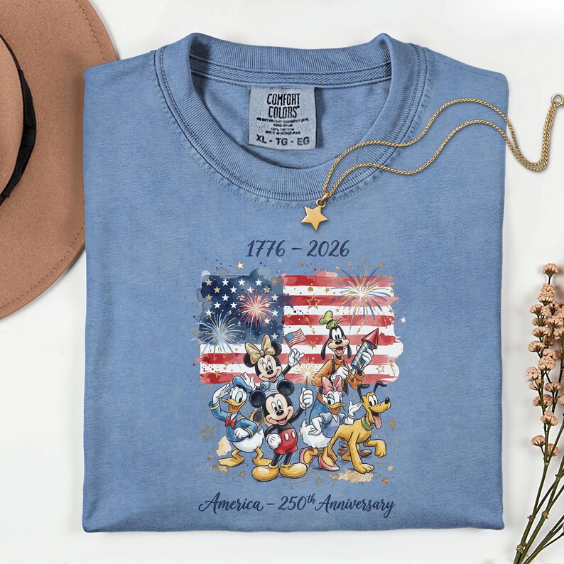 May include: A light blue Comfort Colors t-shirt featuring a graphic of the American flag with fireworks and Disney characters. The text reads "1776-2026" and "America - 250th Anniversary". A gold star pendant necklace rests on the shirt.