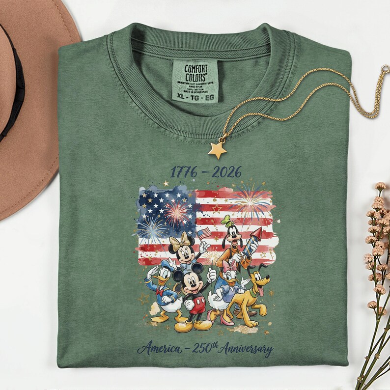 May include: Green XL t-shirt featuring a colorful graphic of Disney characters in front of an American flag with fireworks. The text reads "1776-2026" and "America - 250th Anniversary". A gold star necklace is draped on the shirt.