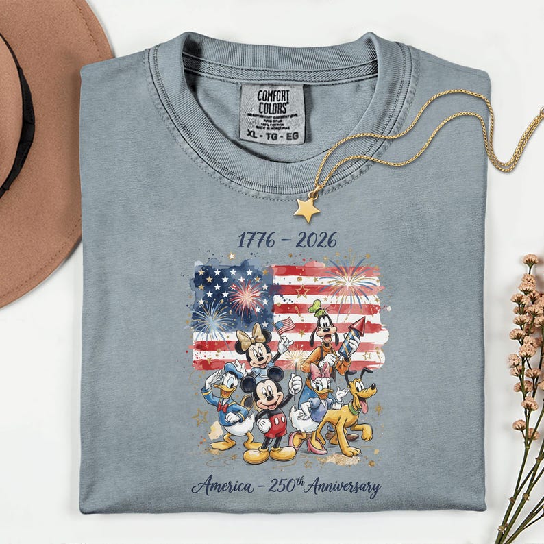 May include: A light blue t-shirt featuring a colorful graphic of Disney characters in front of an American flag with fireworks. The shirt has the text "1776-2026" and "America - 250th Anniversary". A gold necklace with a star pendant is draped on the shirt.