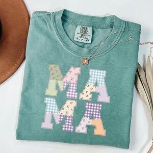 May include: A teal Comfort Colors t-shirt with the word "MAMA" in patchwork letters. The letters feature floral, gingham, and solid patterns in pastel colours. A gold necklace with a pendant rests on the shirt.