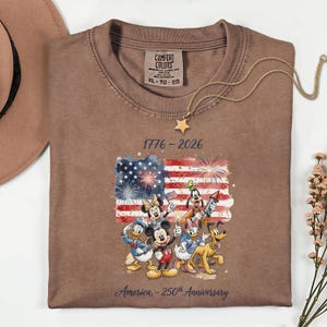 May include: Brown t-shirt with a graphic of the American flag and Disney characters. The shirt has the text "1776-2026" and "America - 250th Anniversary". A gold star necklace is draped over the shirt.