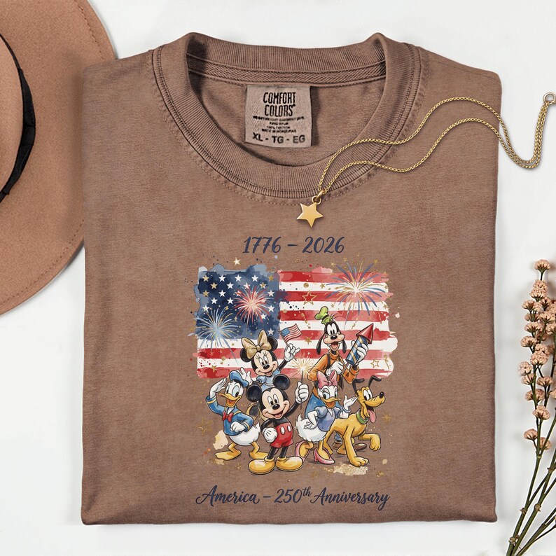 May include: Brown t-shirt with a graphic of the American flag and Disney characters. The shirt has the text "1776-2026" and "America - 250th Anniversary". A gold star necklace is draped over the shirt.