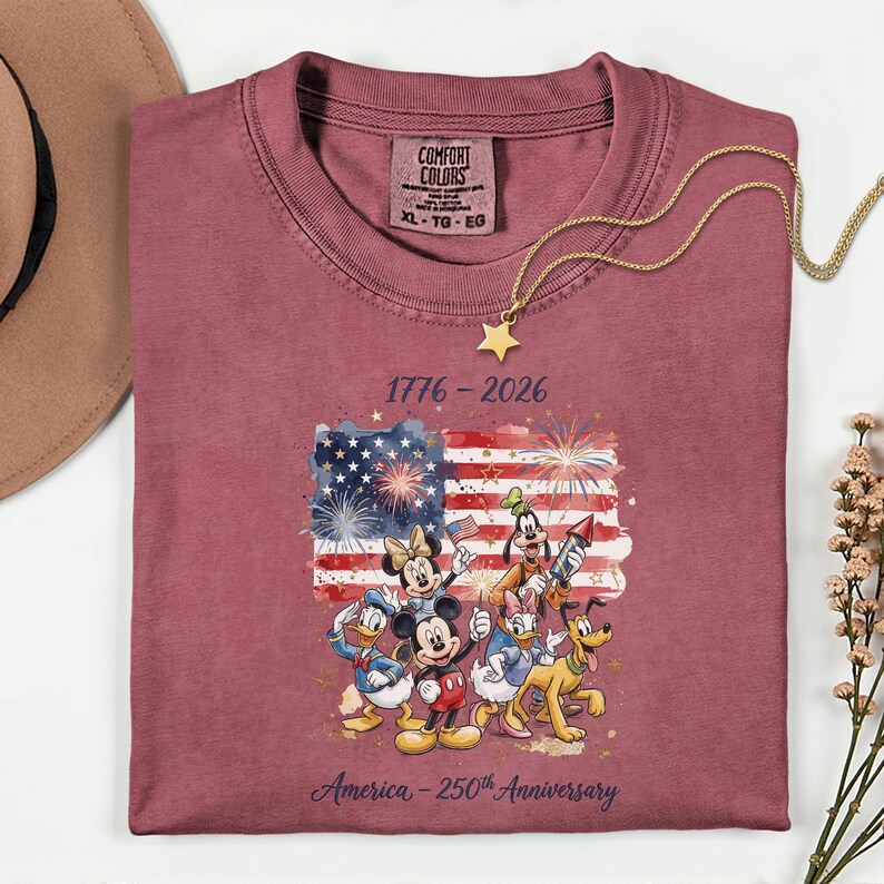 May include: A maroon t-shirt featuring a graphic of the American flag with fireworks and Disney characters. The text "1776-2026" and "America - 250th Anniversary" are printed on the shirt. A gold necklace with a star pendant is draped on the shirt.