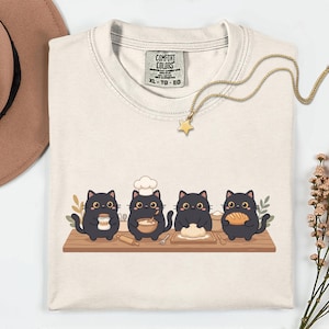 May include: Cream-colored Comfort Colors t-shirt featuring a graphic of four black cats baking bread. The cats are on a wooden surface with flour, a rolling pin, and a loaf of bread. A gold necklace with a star pendant is visible.