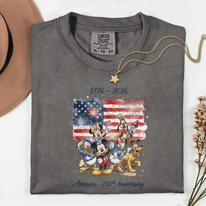May include: A gray Comfort Colors t-shirt featuring a colorful graphic of Disney characters in front of an American flag with fireworks. The text reads "1776-2026" and "America - 250th Anniversary". A gold necklace with a star pendant is draped on the shirt.