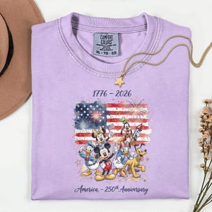 May include: A lilac-colored t-shirt featuring a graphic of the American flag with fireworks and Disney characters. The text reads "1776-2026" and "America - 250th Anniversary". A gold necklace with a star pendant is draped on the shirt.