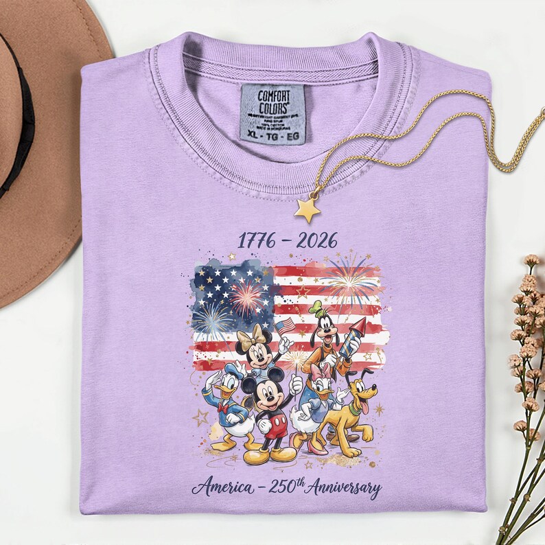 May include: A lilac-colored t-shirt featuring a graphic of the American flag with fireworks and Disney characters. The text reads "1776-2026" and "America - 250th Anniversary". A gold necklace with a star pendant is draped on the shirt.