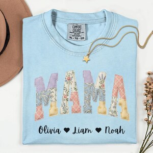 Puede incluir: Camiseta azul claro con la palabra "MAMA" en letras de patchwork. Las letras están hechas de varias telas florales y estampadas. Debajo de la palabra están los nombres Olivia, Liam y Noah. Un collar dorado con un colgante de estrella está en la camiseta.