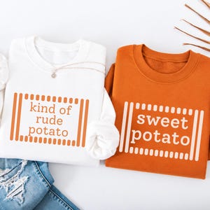 May include: A white sweatshirt with orange text reading kind of rude potato and an orange sweatshirt with white text reading sweet potato. Both garments feature a rectangular border design made of vertical lines and dots surrounding the text.